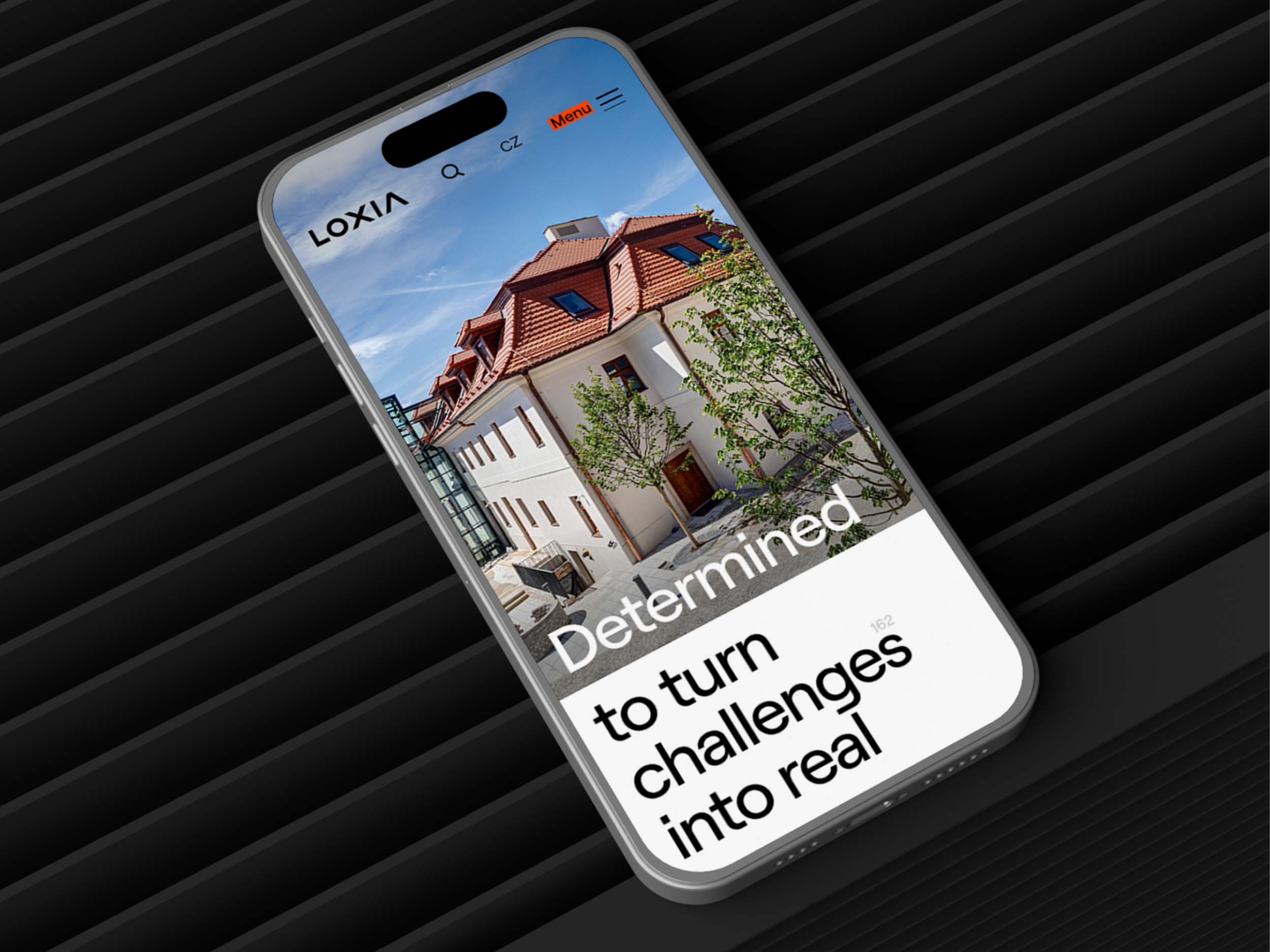Smartphone zobrazuje webovou stránku s nápisem Determined to turn challenges into real na pozadí moderní budovy zapuštěné ve stromech v městském prostředí.