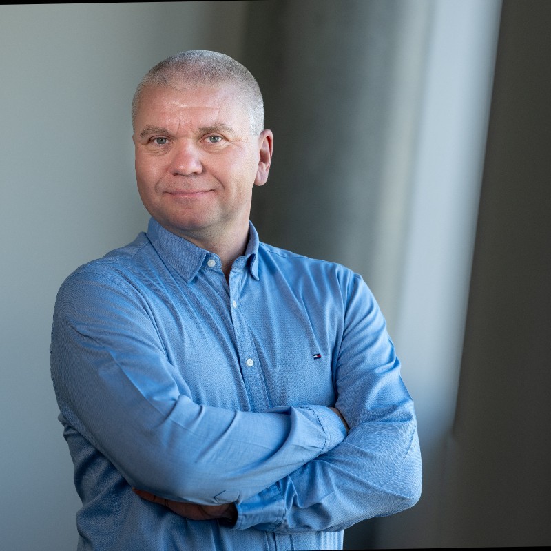 Robert Paskovský, muž s krátkými vlasy ve světle modré košili na knoflíky, stojí se zkříženýma rukama a lehce se usmívá před rozmazaným pozadím interiéru, jemně osvětleným přirozeným světlem.