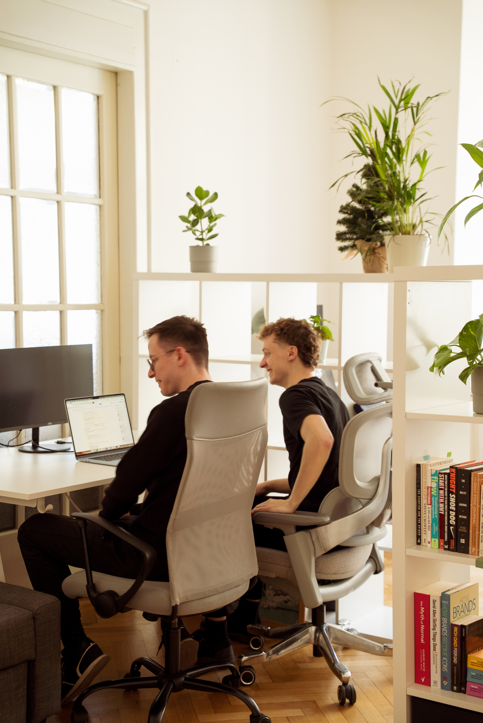 Two men are working in a modern office with light-colored furnishings and green plants. They are sitting in comfortable chairs at a desk with computers and a shelf full of books.