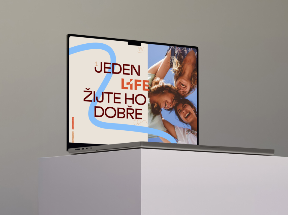 Moderní notebook na stole zobrazující obrazovku s textem Jeden Life Žijte ho dobře a usmívajícími se lidmi. Elegantní design a motivující poselství životního stylu.