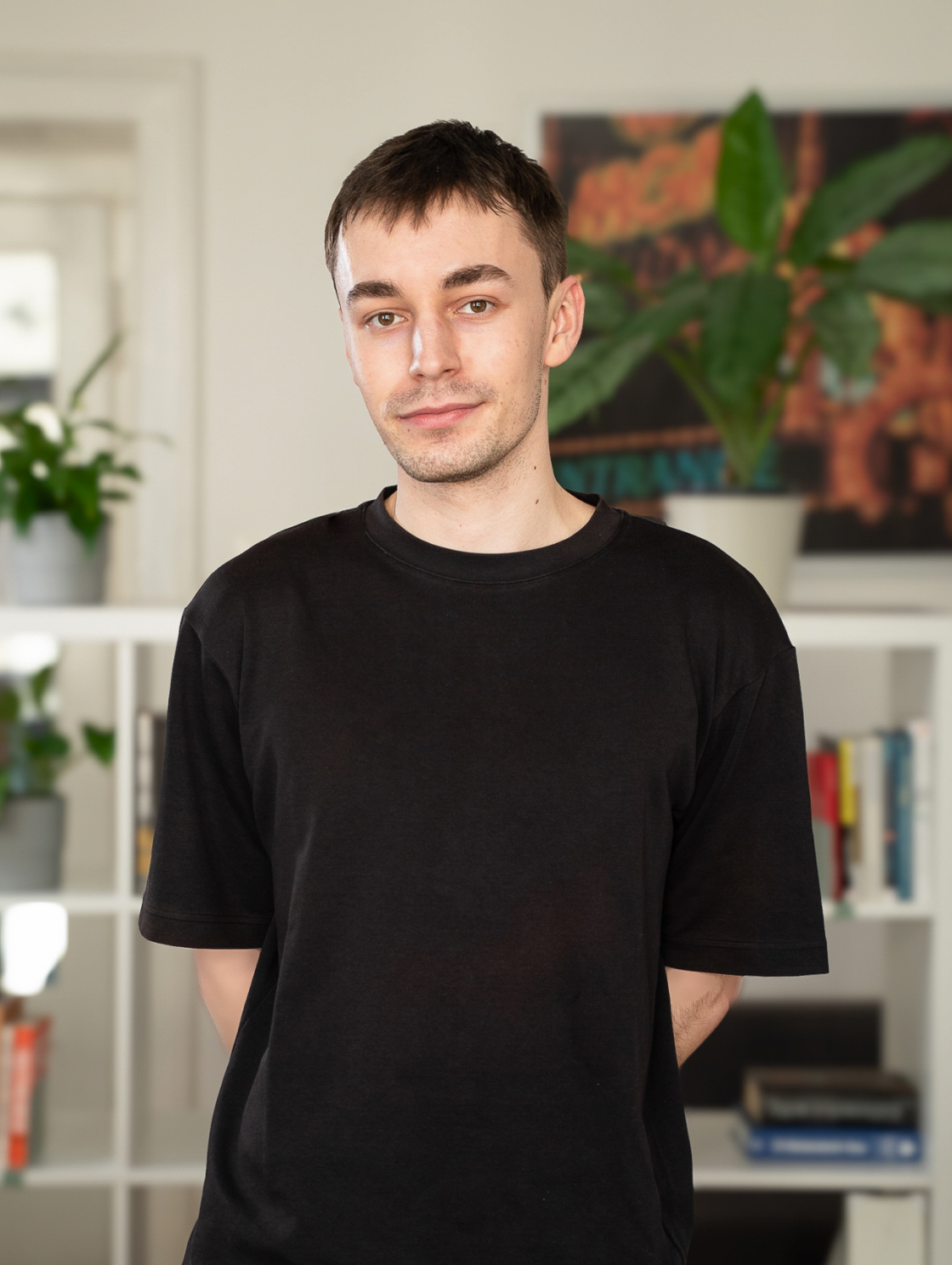 A man in a black T-shirt is standing in front of white shelves filled with books and plants. The setting is modern and fresh, perfect for working or studying at home.