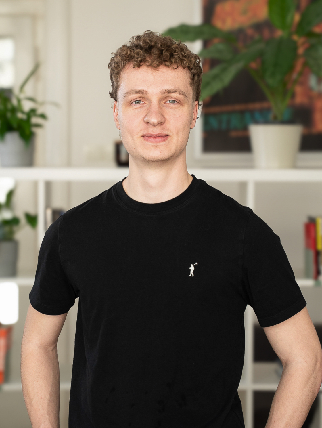 A young man with curly hair, wearing a black T-shirt, is standing in a modern office setting with white shelves and green potted plants in the background.