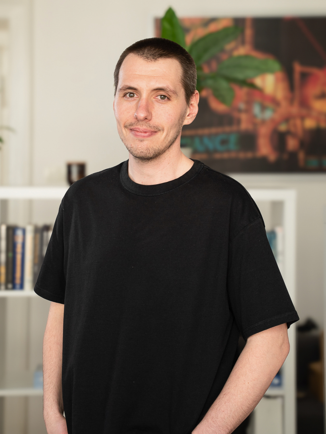 A man in a black T-shirt stands in a modern office with a bookshelf in the background and a green plant next to a painting on the wall; he has a smiling, relaxed expression