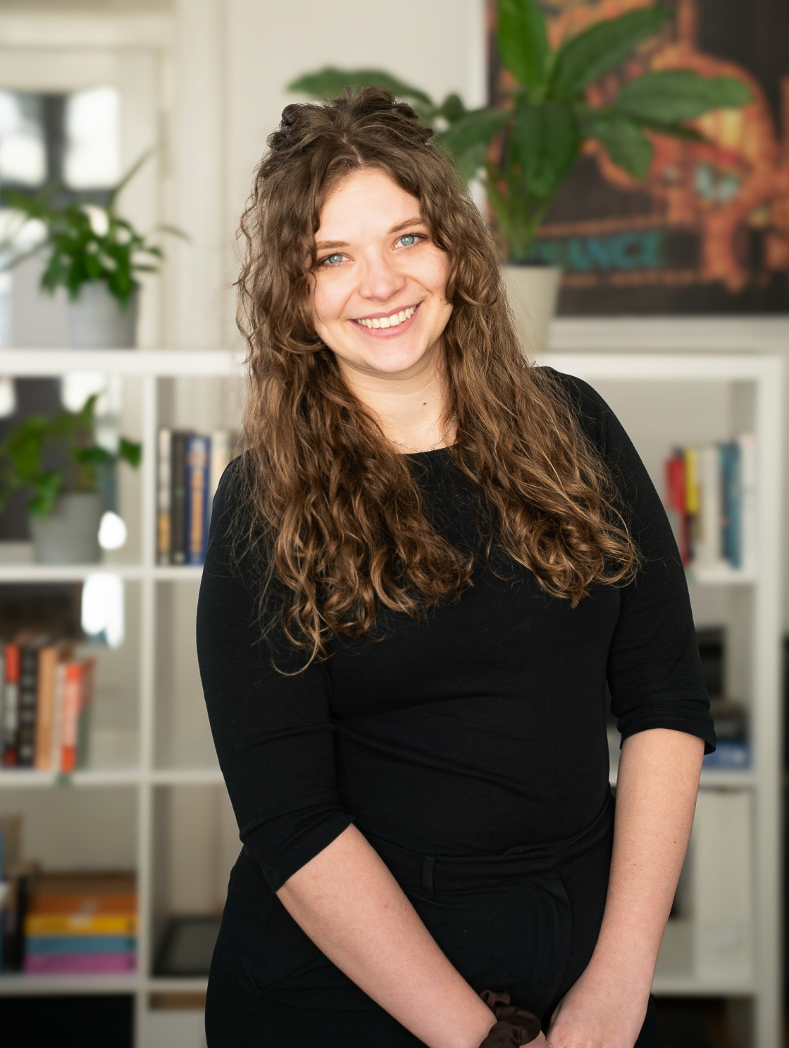 A smiling woman dressed in black is standing in front of a bookshelf. She has long, curly hair, and a green plant and a colorful poster can be seen in the background.