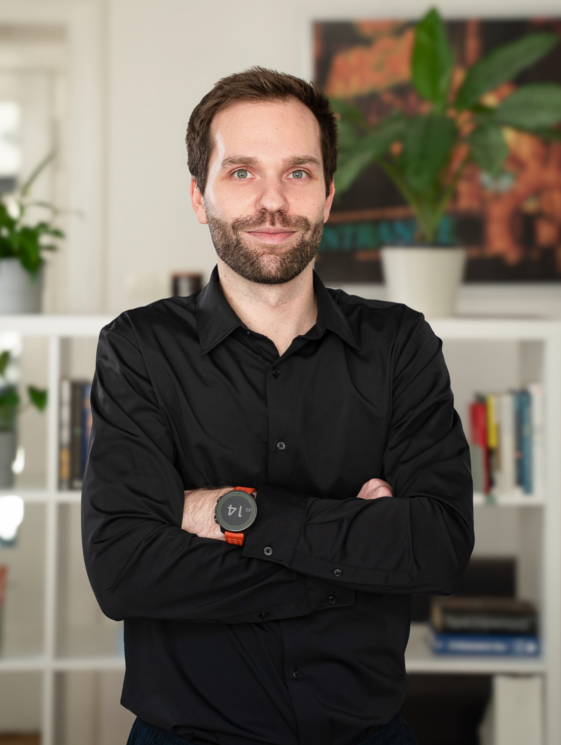 A man in formal black attire is standing in front of a bookshelf with plants in a modern apartment. He has short hair, a beard, and a watch on his left wrist.