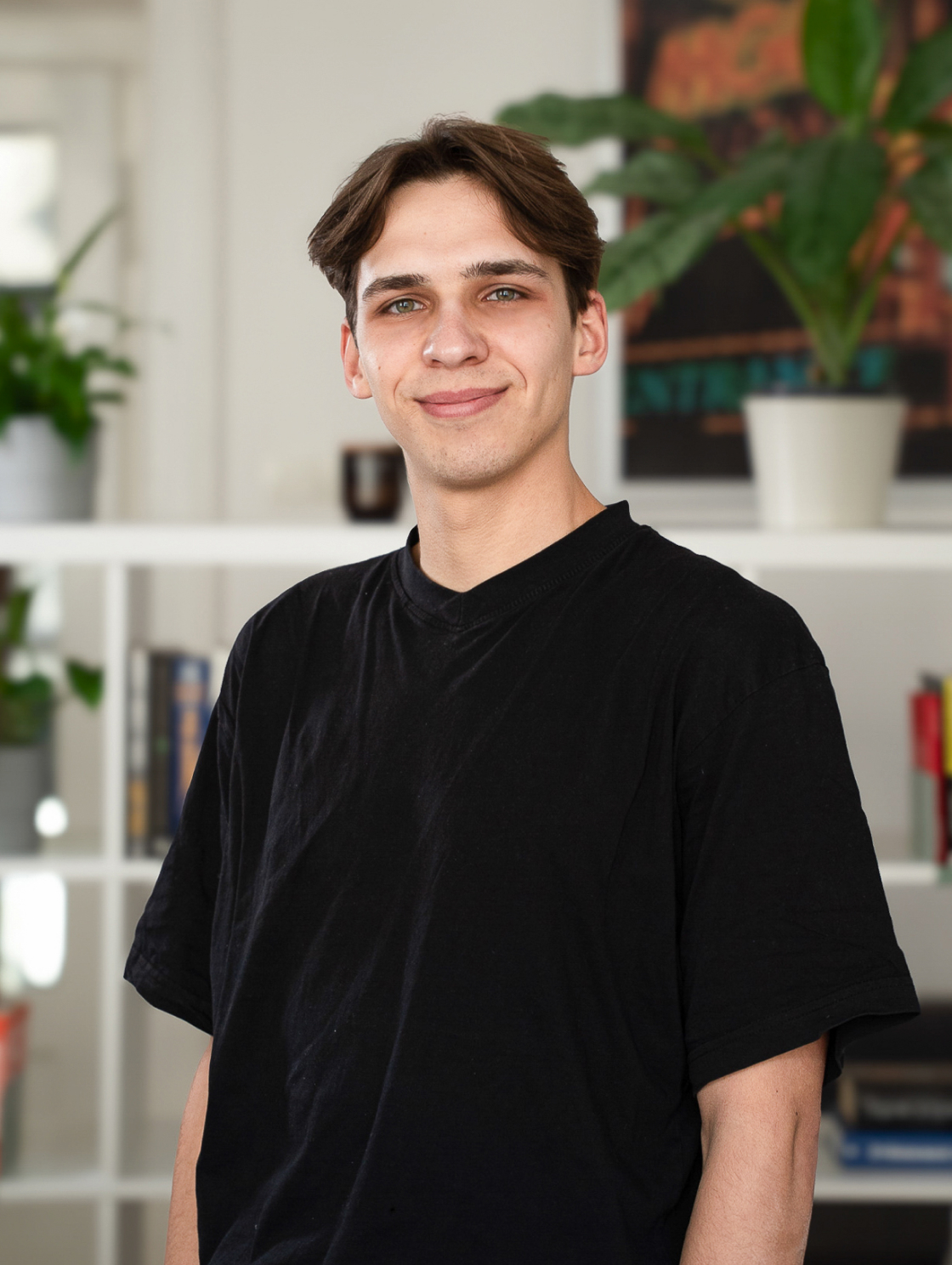 A young man with brown hair is standing in a modern living room with books and plants on the shelves. He is wearing a black T-shirt and smiling slightly at the camera.