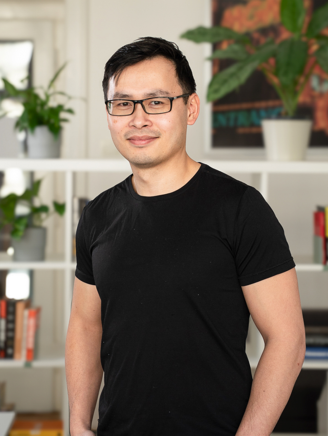 A man in a black T-shirt and glasses is standing in front of a bookshelf with plants. He is smiling and has short black hair. The bright interior gives the picture a welcoming atmosphere.