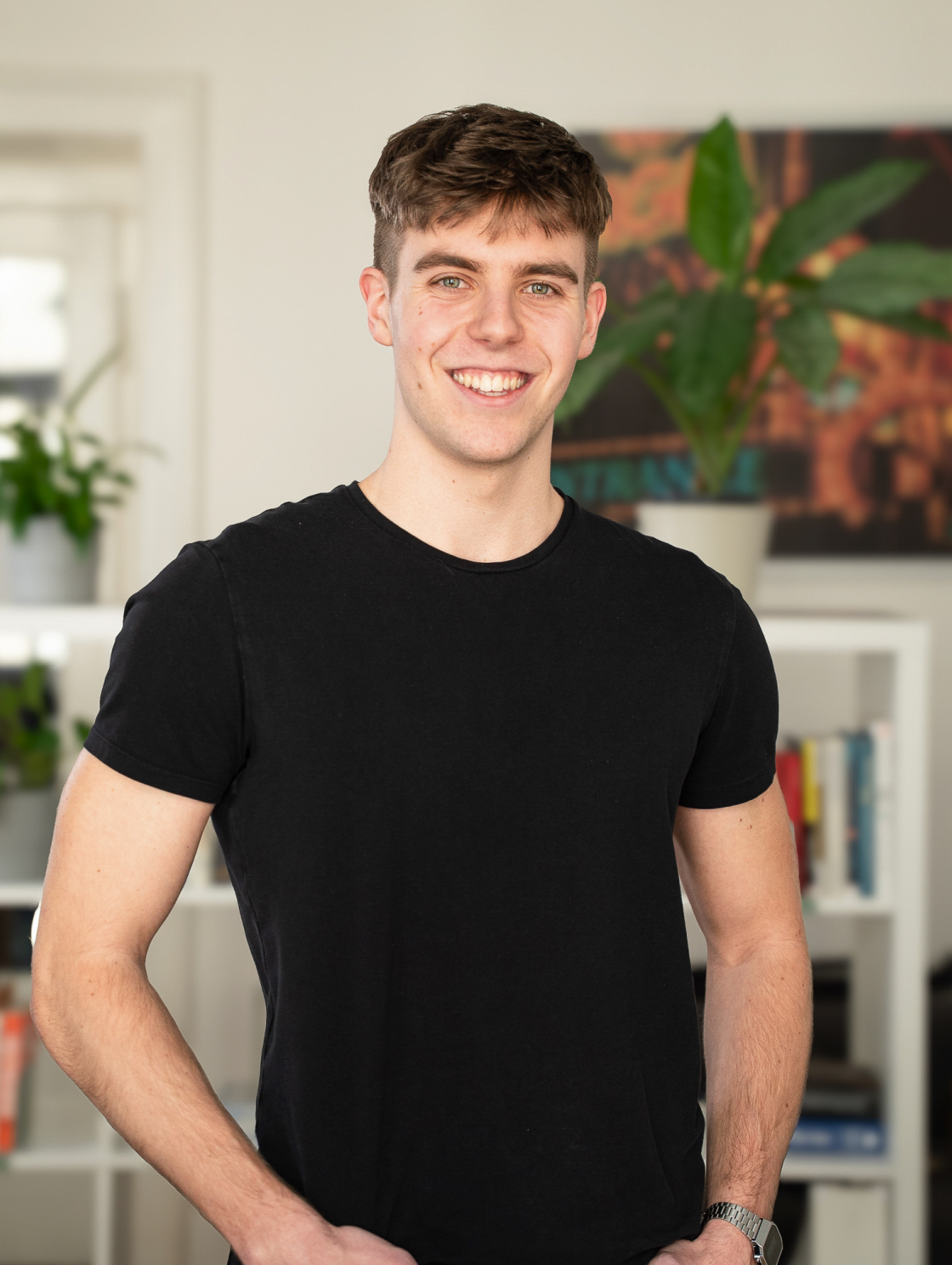 A young man with short hair, wearing a black T-shirt, is standing in a modern, brightly lit room with books and plants in the background, smiling at the camera.