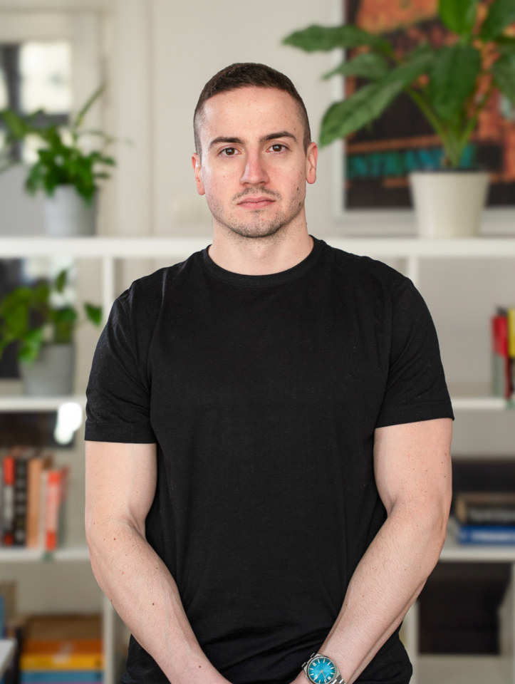 A man wearing a black sports T-shirt stands in front of white shelves filled with books and plants in a modern apartment, well-lit by natural light and ready for the presentation.