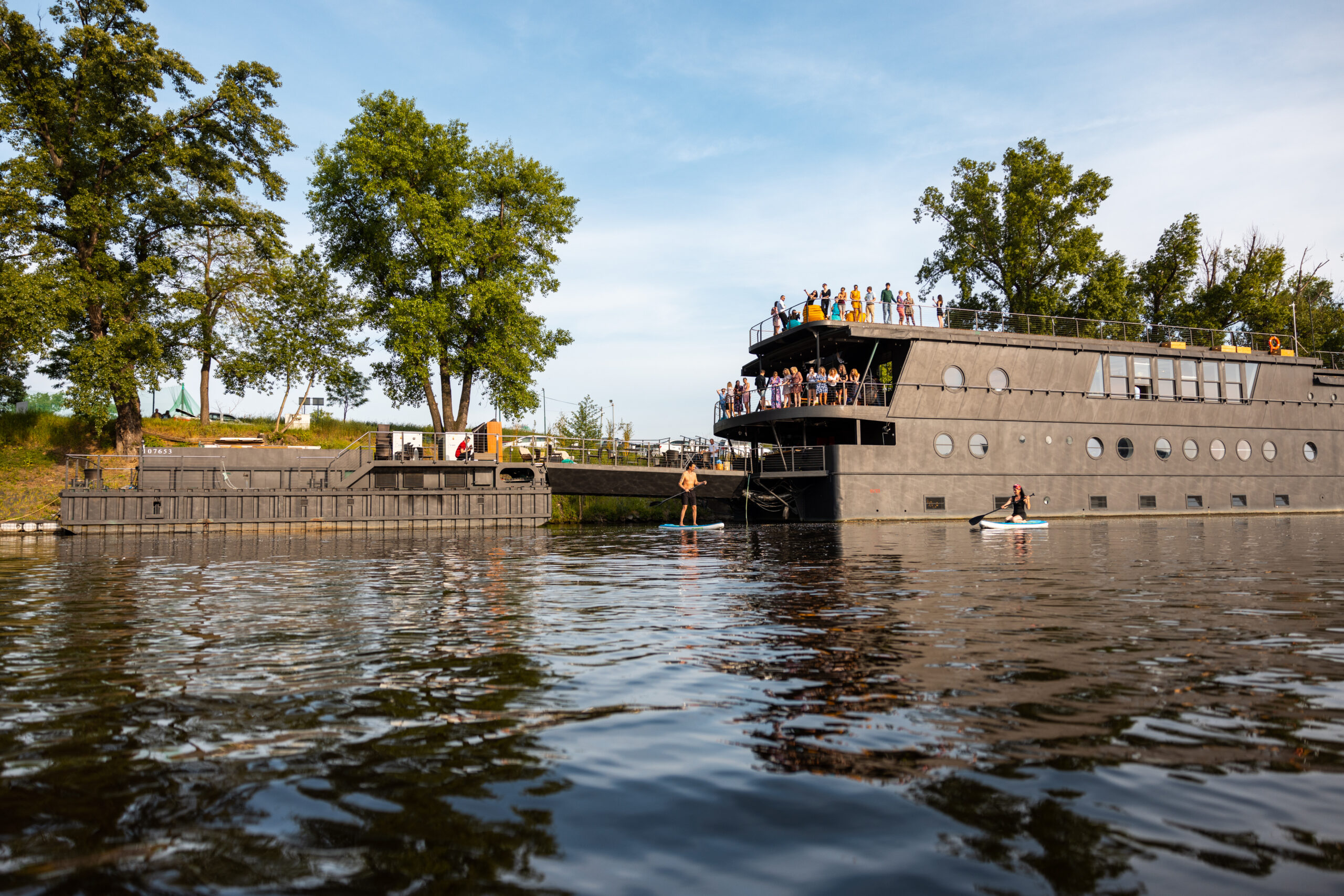 U břehu poblíž stromů kotví moderní tmavě šedý hausbót s lidmi na palubě. V popředí je na vodě osoba na paddleboardu a další v malém kajaku.