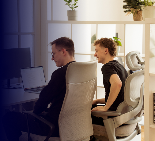 Two men are sitting at a computer in a modern office with light-colored walls and plants. The atmosphere is relaxed and focused on collaboration as they work on a project.