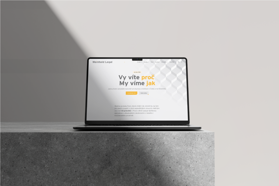 Laptop na betonovém stole s webovou stránkou zobrazenou na obrazovce částečně zastíněný. Moderní design s bílým a šedým pozadím a textem v češtině.