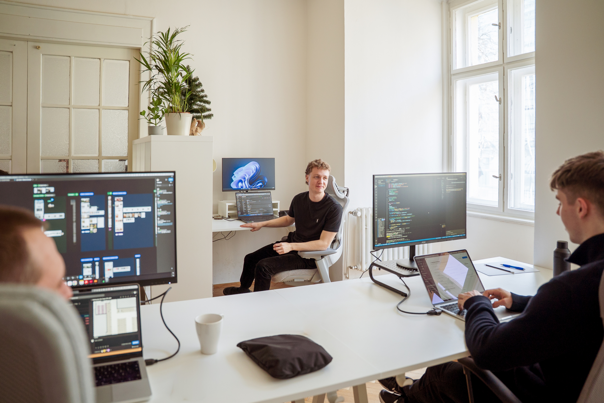 Three men are working on computers in a modern office equipped with large monitors. They foster effective collaboration and communication while developing software in a pleasant environment.