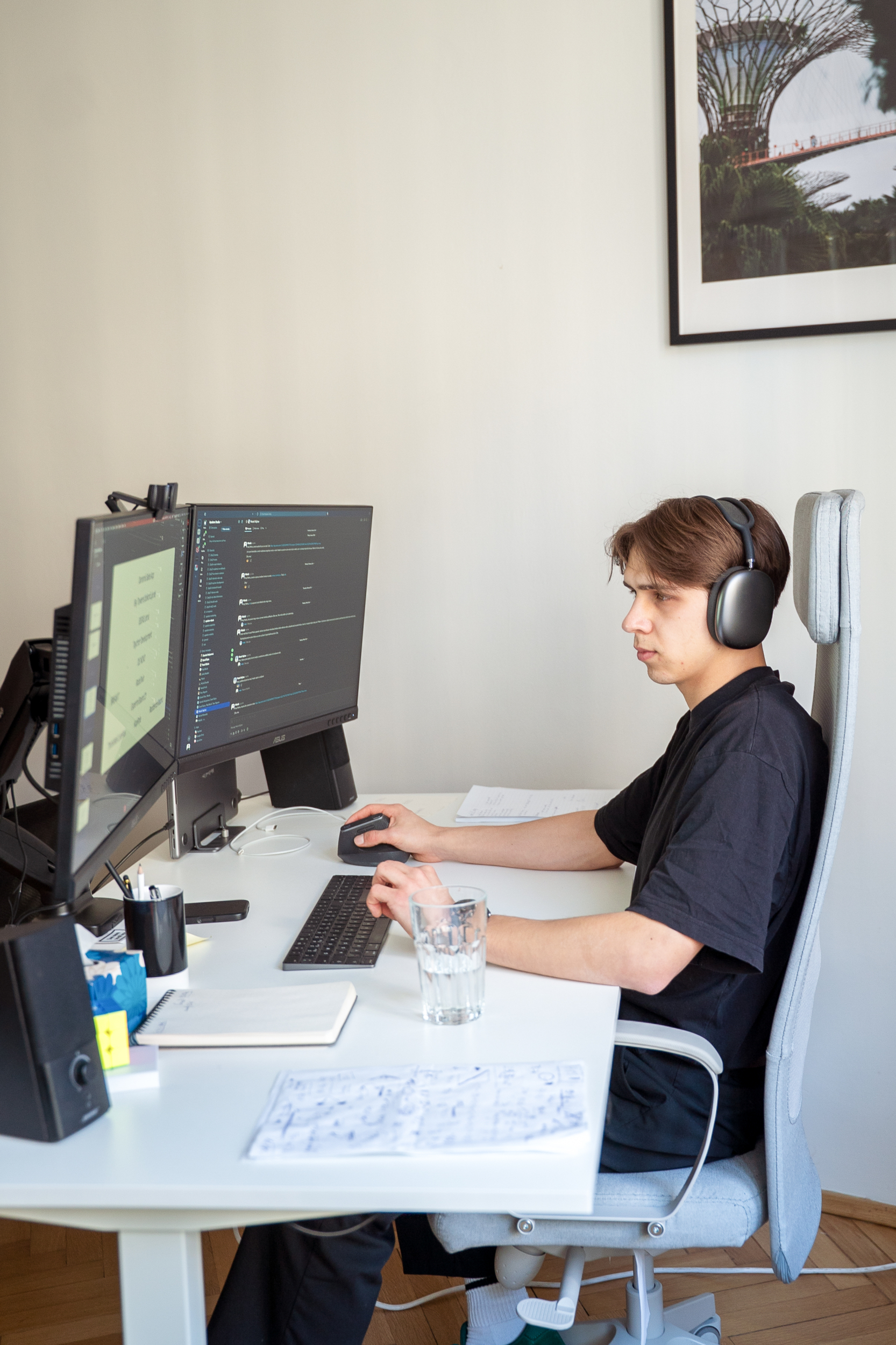 A young man wearing black headphones is working at a desk with two monitors in a modern office—a developer workspace designed to encourage productivity and focus on tasks
