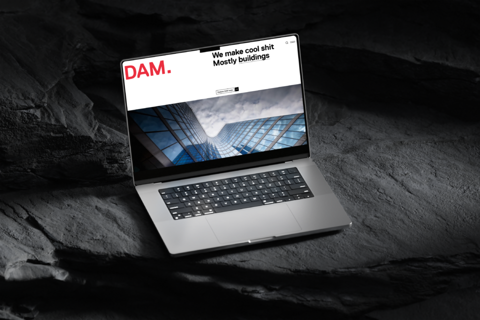 A laptop displaying the DAM Architekti website, the DAM. logo, and a photo of a glass-walled building