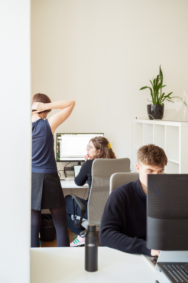 People are working in a modern office with plants on white shelves and two white desks. A man is sitting at a computer monitor, and another woman is stretching behind him.
