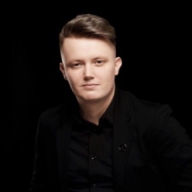 Na tmavém pozadí sedí osoba s krátkými světle hnědými vlasy v černém obleku a černé košili a s lehkým úsměvem se dívá přímo do kamery.