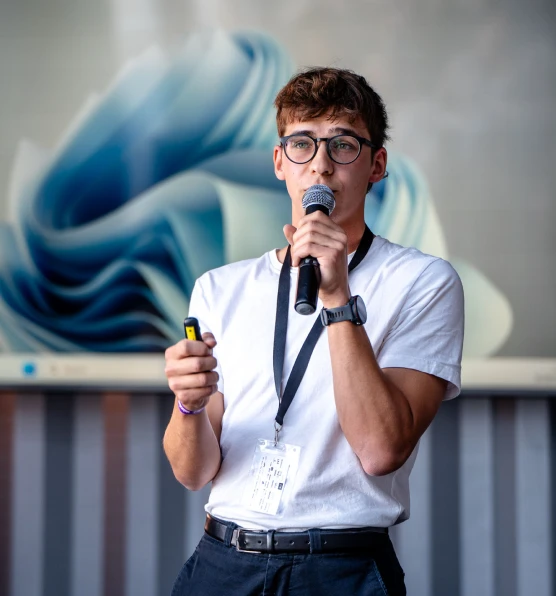 A young man at a conference is speaking into a microphone and holding a remote control in his hand. He is wearing a white shirt and a round badge. Behind him is a blurred colorful decoration.